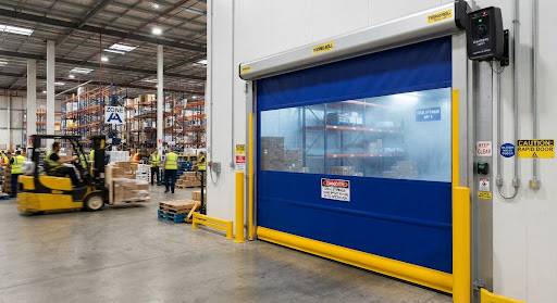Blue fabric high-speed roll-up door with yellow safety guides between a cold storage bay and a warehouse floor with forklift traffic